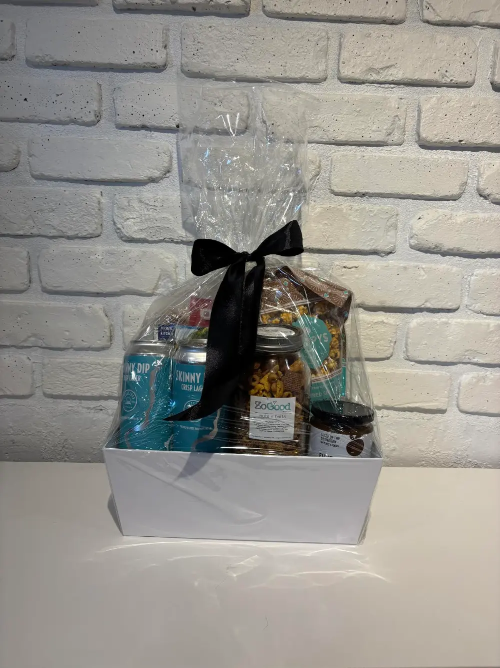 White gift box with two Skinny Dip Crisp Lagers, ZoGood Nuts and Bolts, pear salsa, nacho chips and pepperoni sticks